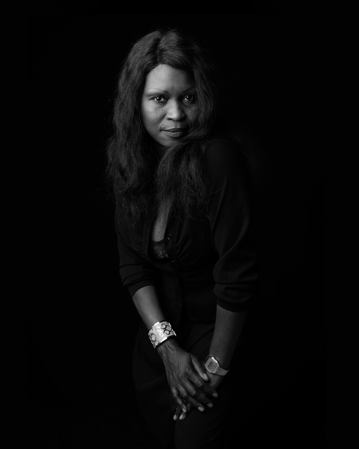 Portrait noir et blanc d’une femme aux cheveux longs, regard intense, mains croisées, fond sombre.