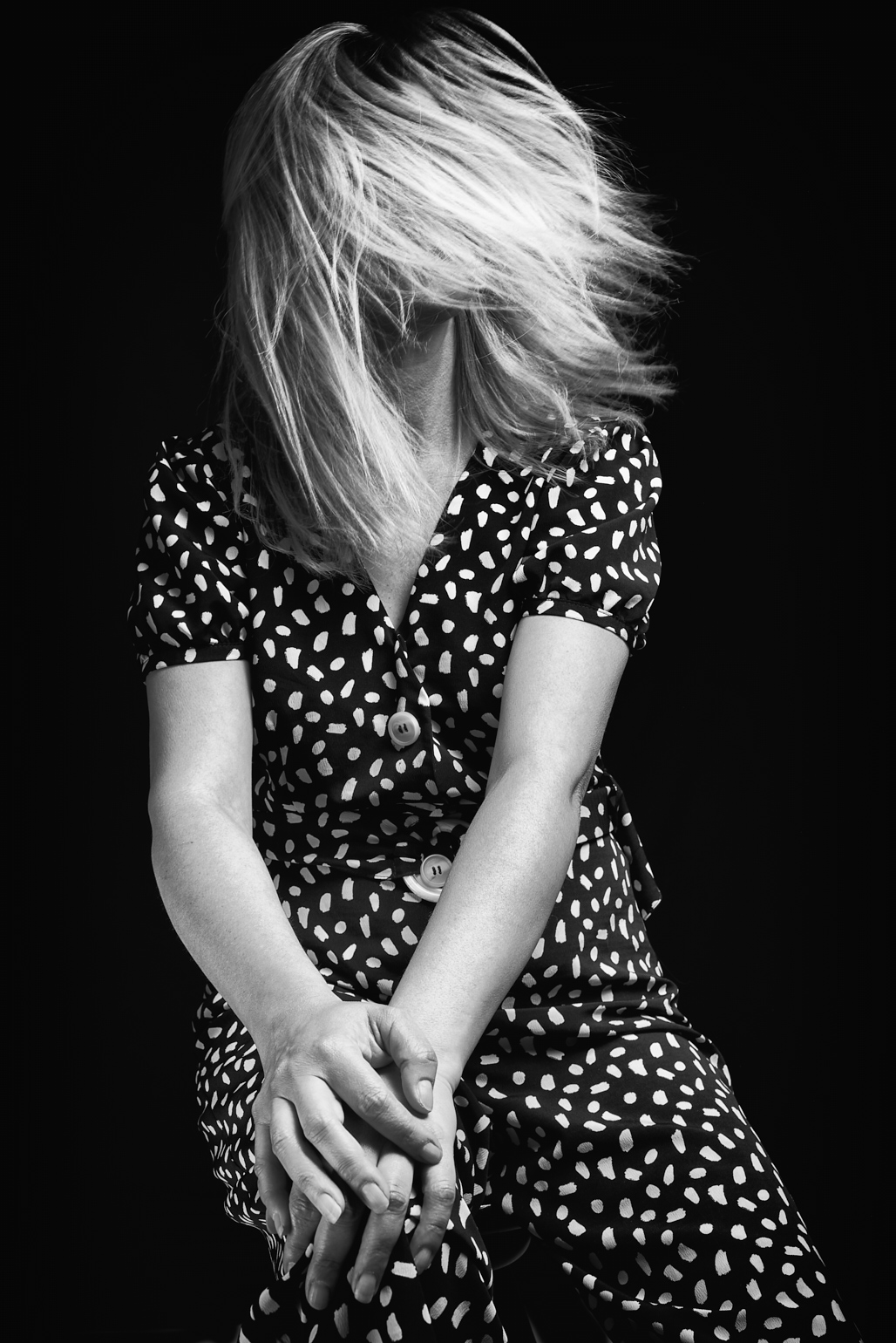 Femme aux cheveux blonds, robe à motifs, mains croisées devant le buste sur fond noir.